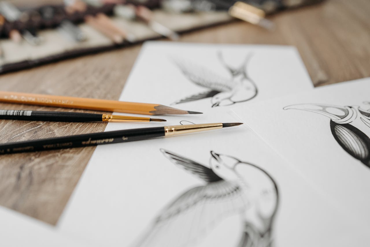 Artistic close-up of bird sketches accompanied by brushes and pencils on a wooden table.