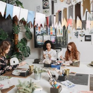 Women engaged in a creative watercolor workshop indoors, surrounded by art supplies and colorful decor.