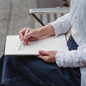 An adult drawing in a sketchbook while sitting outdoors, highlighting creativity and relaxation.